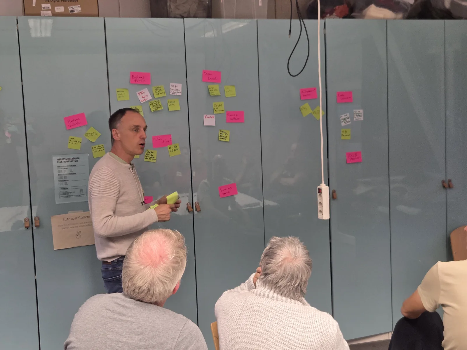 Eine Wand aus Schranktüren, voll beklebt mit pinken und gelben Zetteln; davor steht ein Teilnehmer und erklärt einen Cluster.