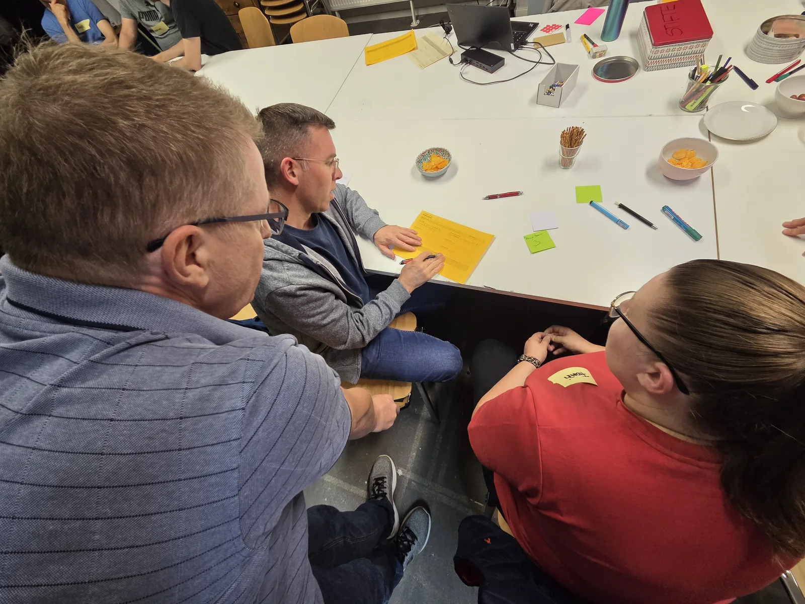 Mehrere Teilnehmende sitzen um einen gelben Projekt-Canvas auf dem Tisch und füllen ihn gemeinsam aus.